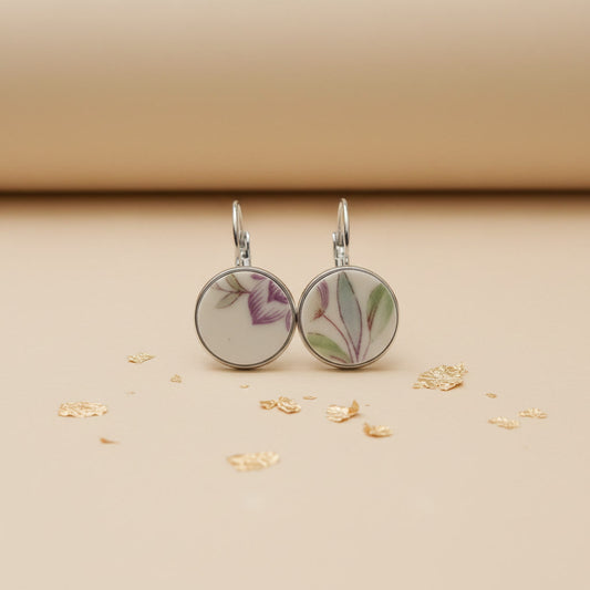 Zwei handgefertigte Porzellan-Ohrhänger des Modells Viola von Edelatelier in silberfarbenen Edelstahlfassungen. Die runden Inlays aus Upcycling-Porzellan zeigen ein dezentes florales Muster mit zart-violetten Blütenkelchen und schmalen, salbeigrünen Blättern. Das Paar ist auf einem beigefarbenen Hintergrund mit dekorativen Goldflocken und sanfter Schattenbildung arrangiert.