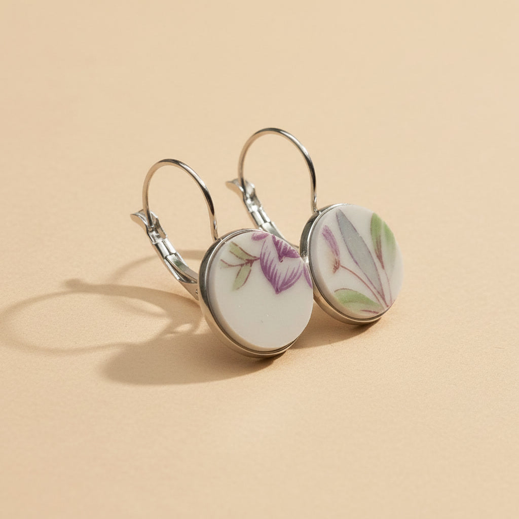Seitenansicht der Viola Porzellan-Ohrringe von Edelatelier, die die hochwertigen Klappbrisuren aus Edelstahl zeigen. Die runden Inlays aus Upcycling-Porzellan präsentieren ein dezentes florales Muster mit zart-violetten Blütenkelchen und schmalen, salbeigrünen Blättern. Das Paar ist auf einem beigefarbenen Hintergrund mit dekorativen Goldflocken und sanfter Schattenbildung arrangiert.