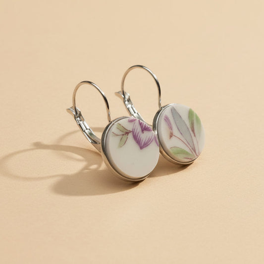 Seitenansicht der Viola Porzellan-Ohrringe von Edelatelier, die die hochwertigen Klappbrisuren aus Edelstahl zeigen. Die runden Inlays aus Upcycling-Porzellan präsentieren ein dezentes florales Muster mit zart-violetten Blütenkelchen und schmalen, salbeigrünen Blättern. Das Paar ist auf einem beigefarbenen Hintergrund mit dekorativen Goldflocken und sanfter Schattenbildung arrangiert.