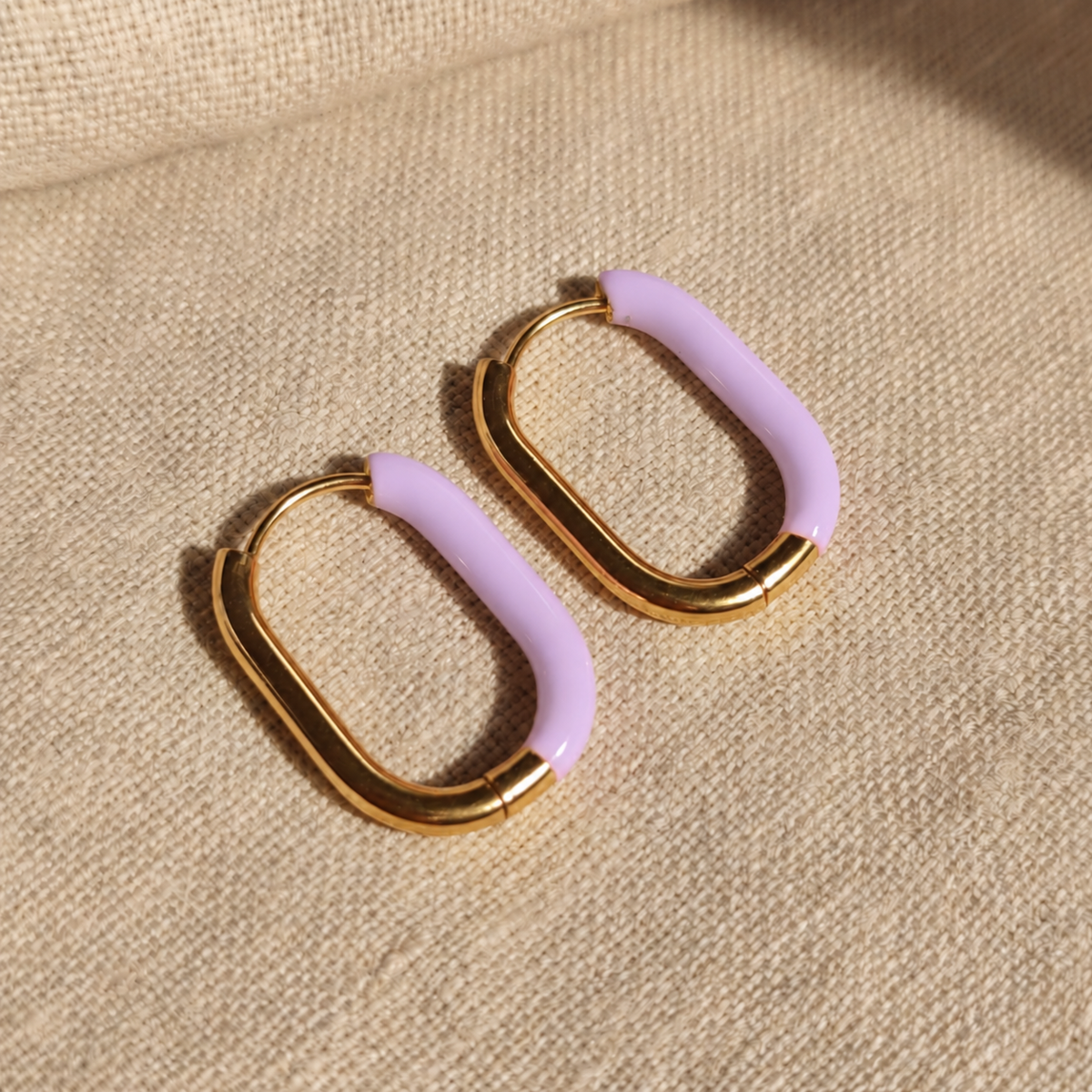 Lavanda Creolen Gold Lavendel frontal auf beigem Leinen, ovale Huggie-Form und lavendelfarbener Akzent sichtbar.