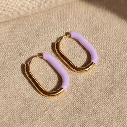 Lavanda Creolen Gold Lavendel frontal auf beigem Leinen, ovale Huggie-Form und lavendelfarbener Akzent sichtbar.
