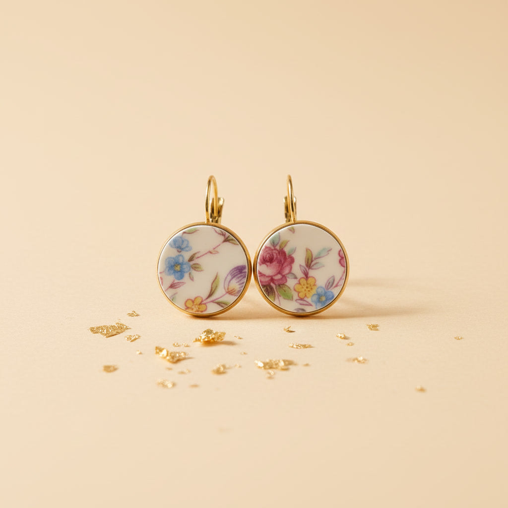 Ein Paar handgefertigte Porzellanohrringe "Millefleurs" von EDELATELIER. auf einem hellen, sandfarbenen Hintergrund mit goldenen Akzenten. Die runden Ohrhänger präsentieren ein farbenfrohes, dichtes Blumendekor mit blauen, rosa und gelben Blüten auf weißem Porzellan, eingefasst in eine goldfarbene Fassung.