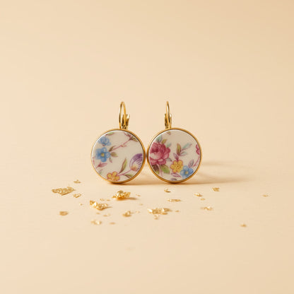 Ein Paar handgefertigte Porzellanohrringe "Millefleurs" von EDELATELIER. auf einem hellen, sandfarbenen Hintergrund mit goldenen Akzenten. Die runden Ohrhänger präsentieren ein farbenfrohes, dichtes Blumendekor mit blauen, rosa und gelben Blüten auf weißem Porzellan, eingefasst in eine goldfarbene Fassung.