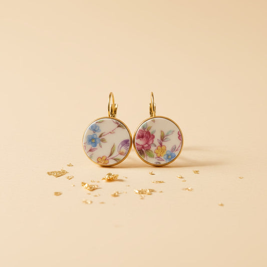 Ein Paar handgefertigte Porzellanohrringe "Millefleurs" von EDELATELIER. auf einem hellen, sandfarbenen Hintergrund mit goldenen Akzenten. Die runden Ohrhänger präsentieren ein farbenfrohes, dichtes Blumendekor mit blauen, rosa und gelben Blüten auf weißem Porzellan, eingefasst in eine goldfarbene Fassung.