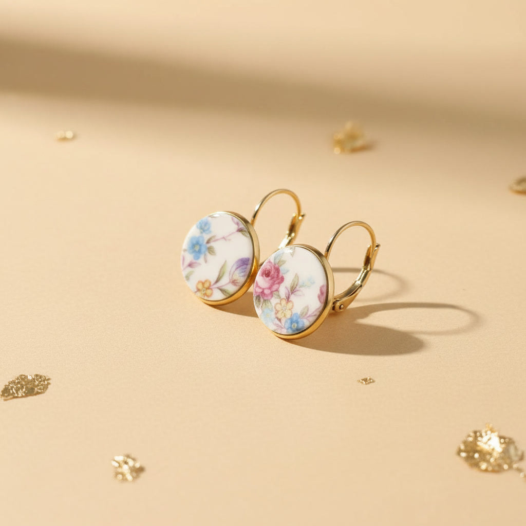 Nahaufnahme der Millefleurs-Ohrringe, die leicht versetzt auf einem beigen Untergrund liegen. Das detaillierte florale Muster und die glänzenden, goldfarbenen Klappbrisuren aus Edelstahl sind deutlich sichtbar und unterstreichen die hochwertige Verarbeitung.