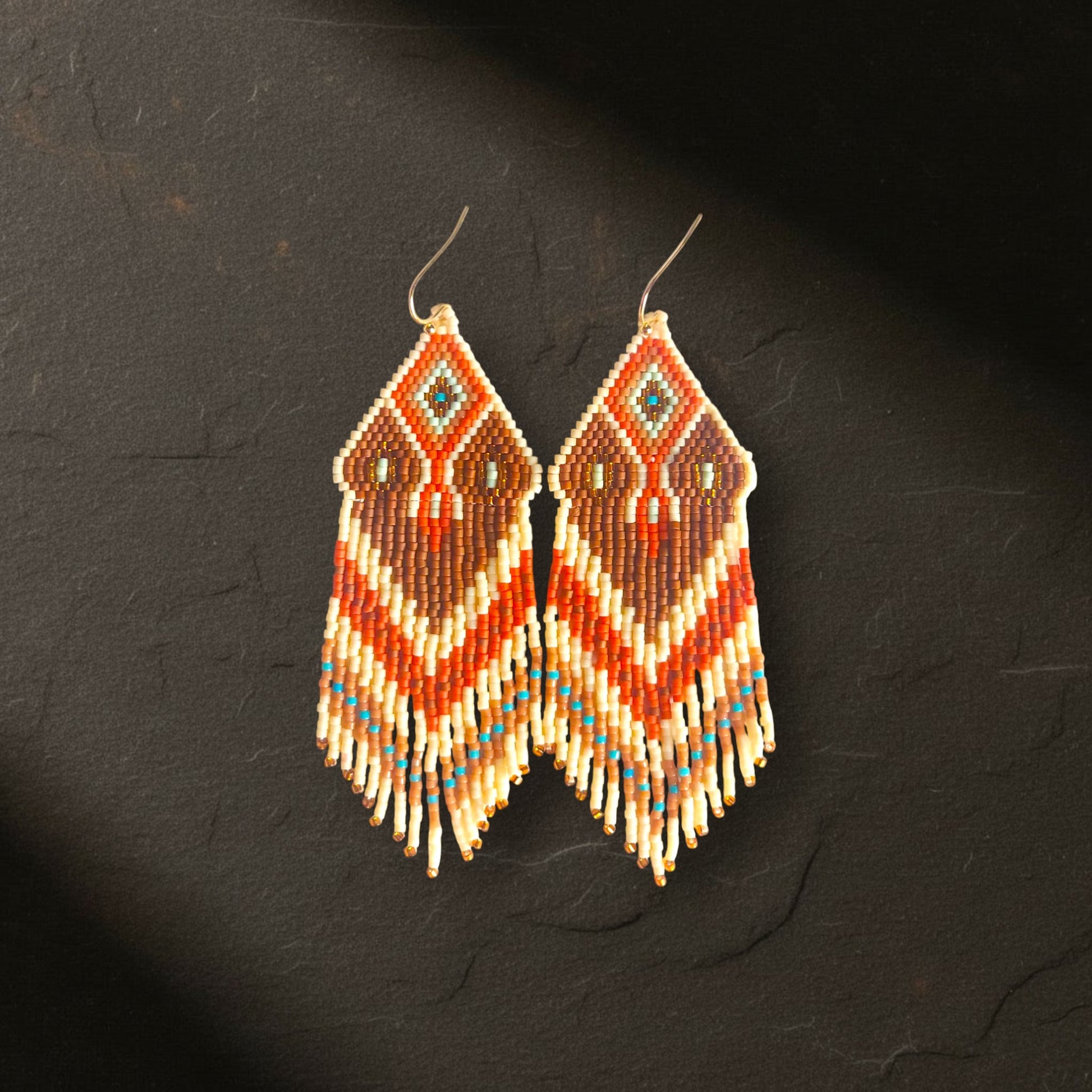 Handgewebte lange Statement-Ohrringe Sabbia Dorata von EDELATELIER. aus Miyuki-Perlen in Creme, Braun und Orange mit türkisfarbenen Akzenten, liegend präsentiert auf einer dunklen Schieferplatte.