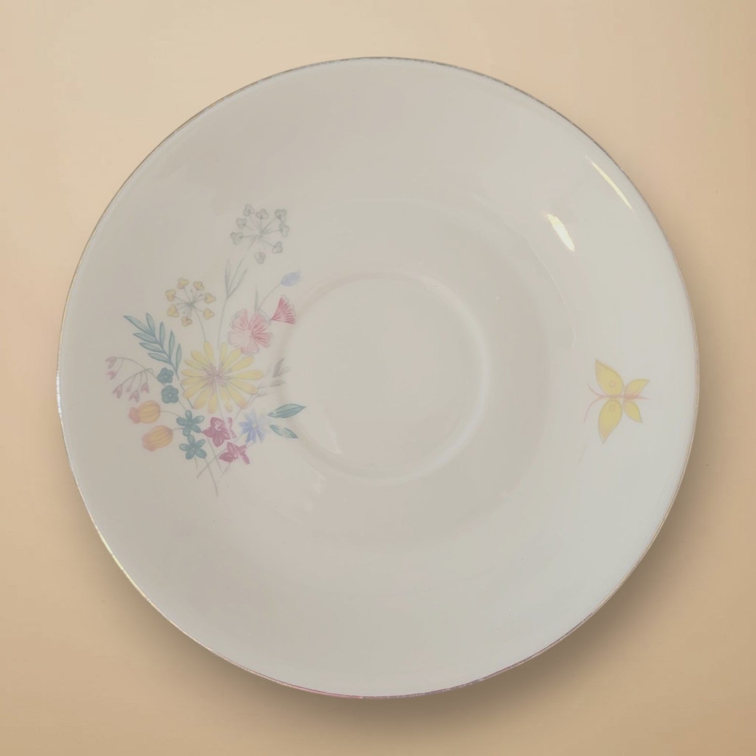 Draufsicht auf die Vorderseite einer weißen Porzellan-Untertasse mit buntem Blumenbouquet links und einem gelben Schmetterling rechts.