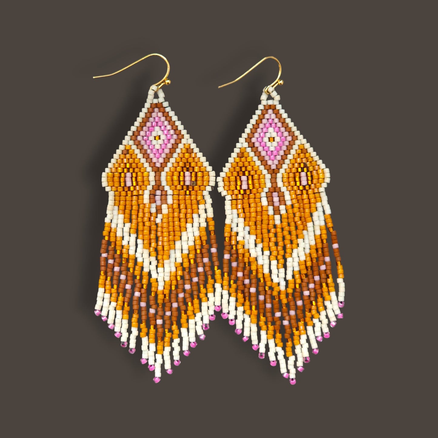 Lange Boho-Ohrhänger aus Miyuki-Perlen mit einem geometrischen Rautenmuster in Weiß, Goldgelb und Rosa sowie langen, farblich abgestuften Fransen an goldfarbenen Ohrhaken vor einem dunklen Hintergrund.