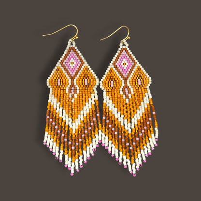 Lange Boho-Ohrhänger aus Miyuki-Perlen mit einem geometrischen Rautenmuster in Weiß, Goldgelb und Rosa sowie langen, farblich abgestuften Fransen an goldfarbenen Ohrhaken vor einem dunklen Hintergrund.
