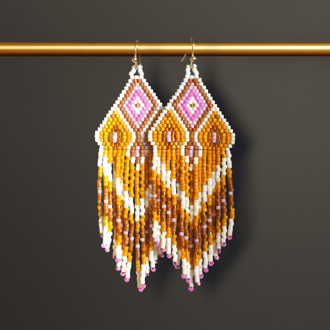 Lange Boho-Ohrhänger aus Miyuki-Perlen mit einem geometrischen Rautenmuster in Weiß, Goldgelb und Rosa sowie langen, farblich abgestuften Fransen an goldfarbenen Ohrhaken an einer goldenen Stange hängend. 