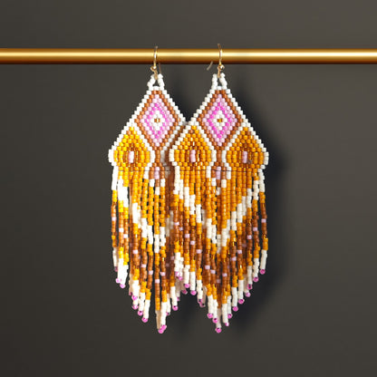 Lange Boho-Ohrhänger aus Miyuki-Perlen mit einem geometrischen Rautenmuster in Weiß, Goldgelb und Rosa sowie langen, farblich abgestuften Fransen an goldfarbenen Ohrhaken an einer goldenen Stange hängend. 