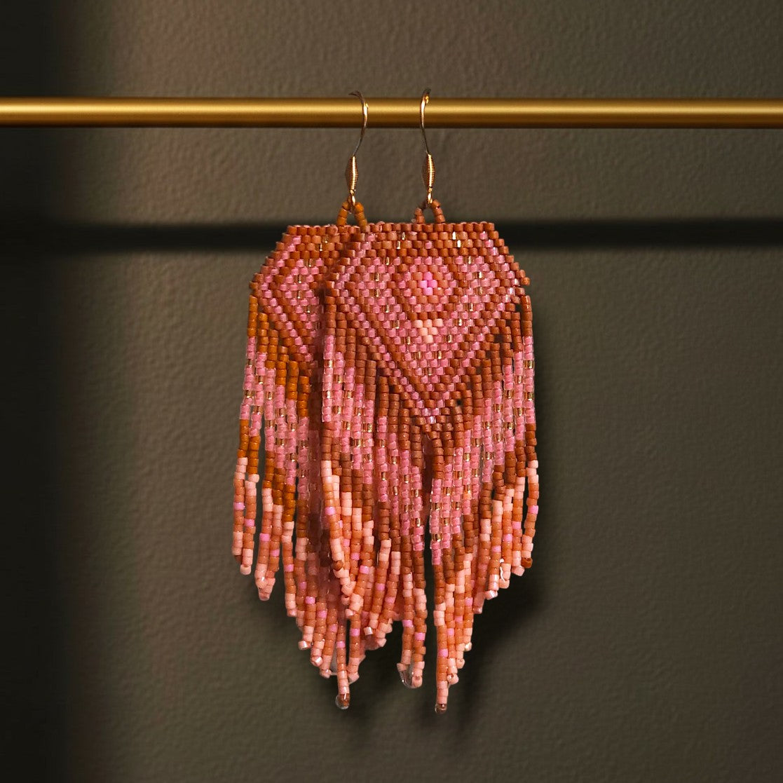 Handgewebte Miyuki Perlenohrringe Alba del Deserto, aufgehängt an einer goldenen Präsentationsstange vor einem dunklen Hintergrund. Das Design zeigt warme Wüstentöne in Rosé, Terrakotta und Apricot mit fließenden Fransen.