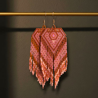 Handgewebte Miyuki Perlenohrringe Alba del Deserto, aufgehängt an einer goldenen Präsentationsstange vor einem dunklen Hintergrund. Das Design zeigt warme Wüstentöne in Rosé, Terrakotta und Apricot mit fließenden Fransen.