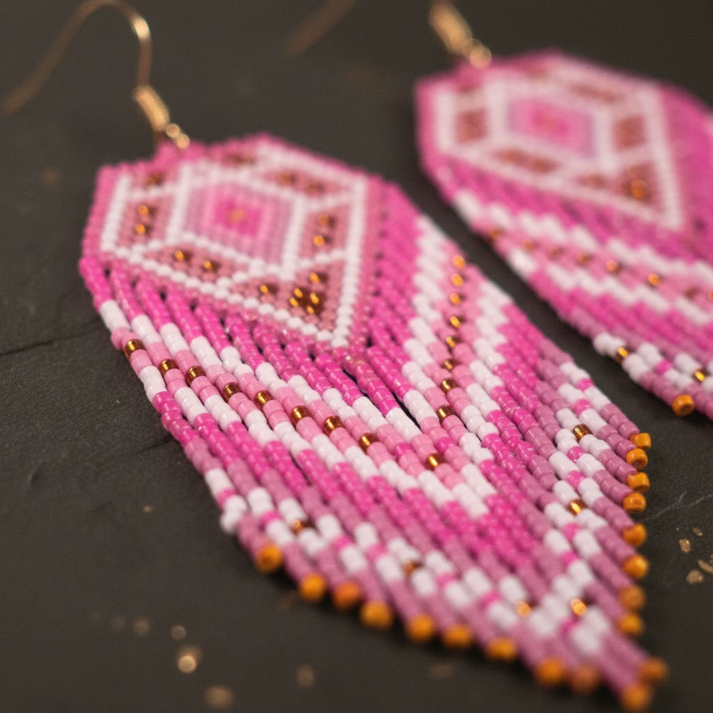 Die pink-goldenen Statement-Ohrringe liegen in einer diagonalen Perspektive flach auf einer dunklen, strukturierten Oberfläche. Der Blickwinkel zeigt die feinen Übergänge der Perlenreihen und die glänzenden goldfarbenen Edelstahl-Ohrhaken.