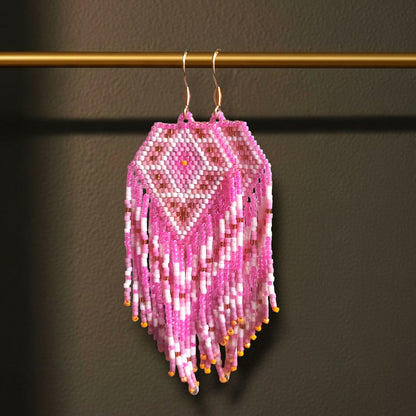 Handgewebte Miyuki Perlenohrringe Alba Dorata, aufgehängt an einer goldenen Präsentationsstange vor einem dunklen Hintergrund. Das Design zeigt ein warmes Rautenmuster in glänzendem Rosé, Pink und Goldtönen mit langen, fließenden Fransen.