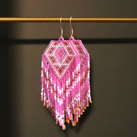 Handgewebte Miyuki Perlenohrringe Alba Dorata, aufgehängt an einer goldenen Präsentationsstange vor einem dunklen Hintergrund. Das Design zeigt ein warmes Rautenmuster in glänzendem Rosé, Pink und Goldtönen mit langen, fließenden Fransen.