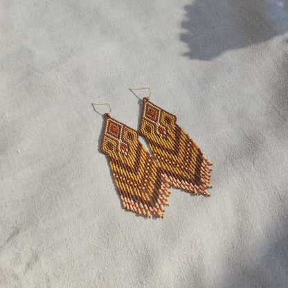 Ambra di Sole Miyuki Ohrringe frontal auf Leinen, langes geometrisches Perlenmuster mit beweglichen Fransen.