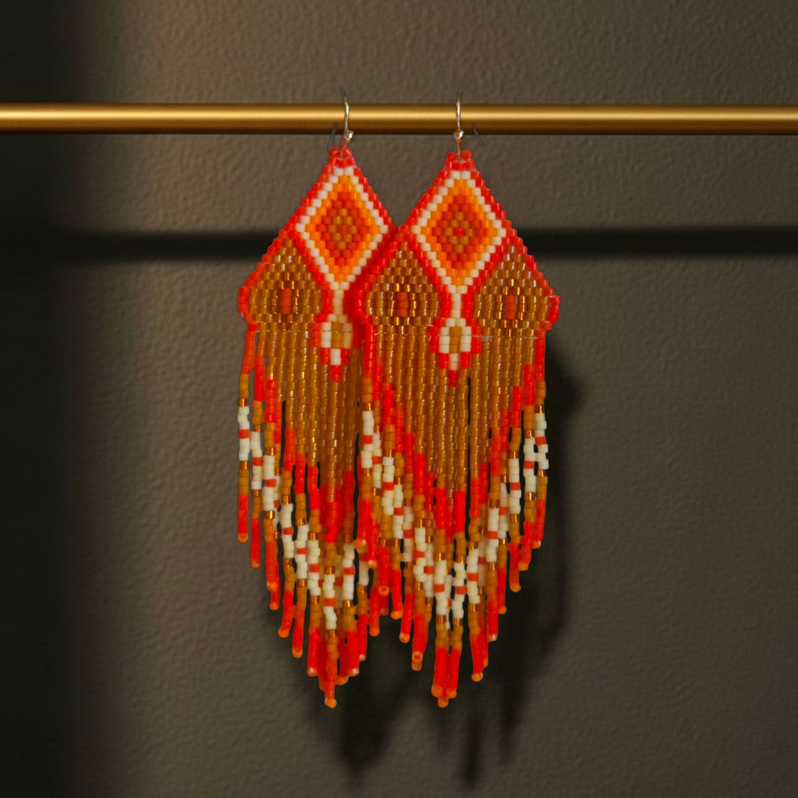 Handgewebte lange Statement-Ohrringe Arancia d'Oro von EDELATELIER. in leuchtendem Orange, Gold und Beige, elegant hängend an einer goldenen Stange vor dunklem Hintergrund.