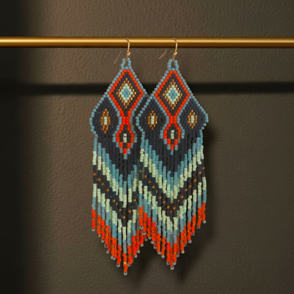 Lange Boho-Ohrhänger aus Miyuki-Perlen mit einem markanten Rautenmuster in Anthrazit, Hellblau und kräftigem Orange sowie langen, farblich abgestimmten Fransen an goldfarbenen Ohrhaken an einer goldenen Stange. 