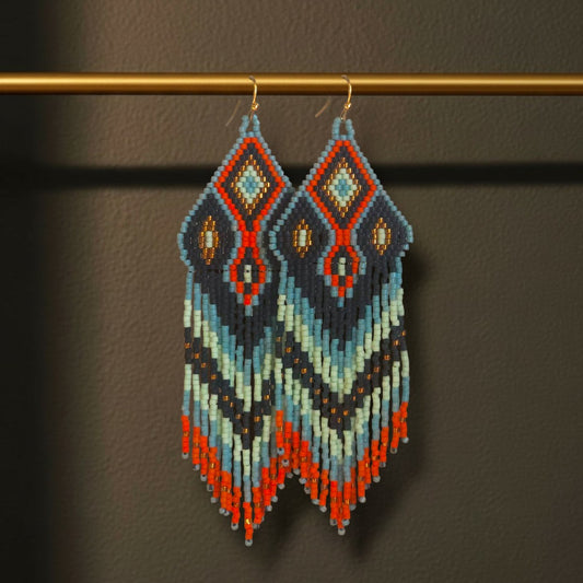 Lange Boho-Ohrhänger aus Miyuki-Perlen mit einem markanten Rautenmuster in Anthrazit, Hellblau und kräftigem Orange sowie langen, farblich abgestimmten Fransen an goldfarbenen Ohrhaken an einer goldenen Stange. 