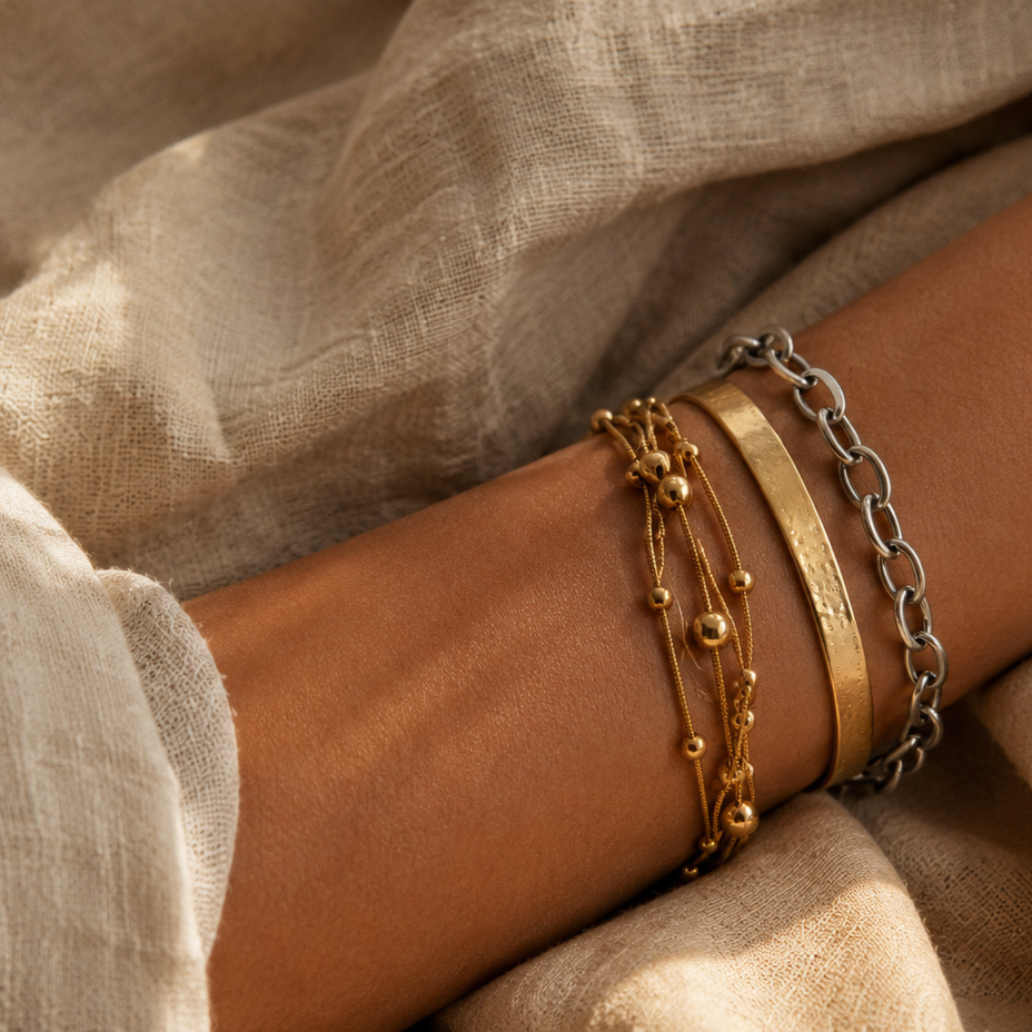 Gold- und silberfarbene Edelstahlarmbänder im Layering-Look am Handgelenk auf hellem Leinen.