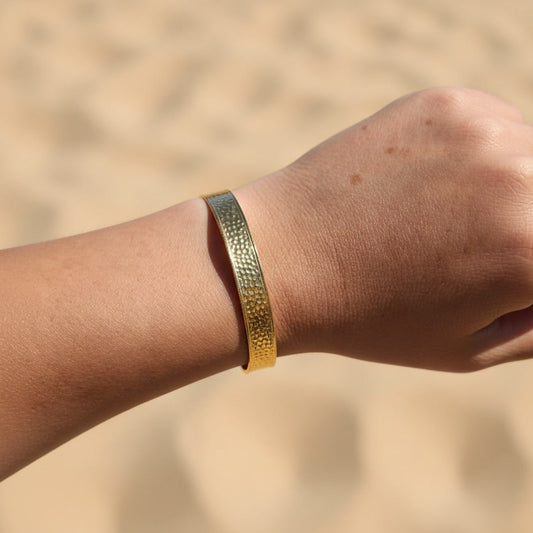 Aurelia Armreif Gold am Handgelenk getragen, breiter gehämmerter Edelstahl-Armreif in 18 Karat PVD Gold.