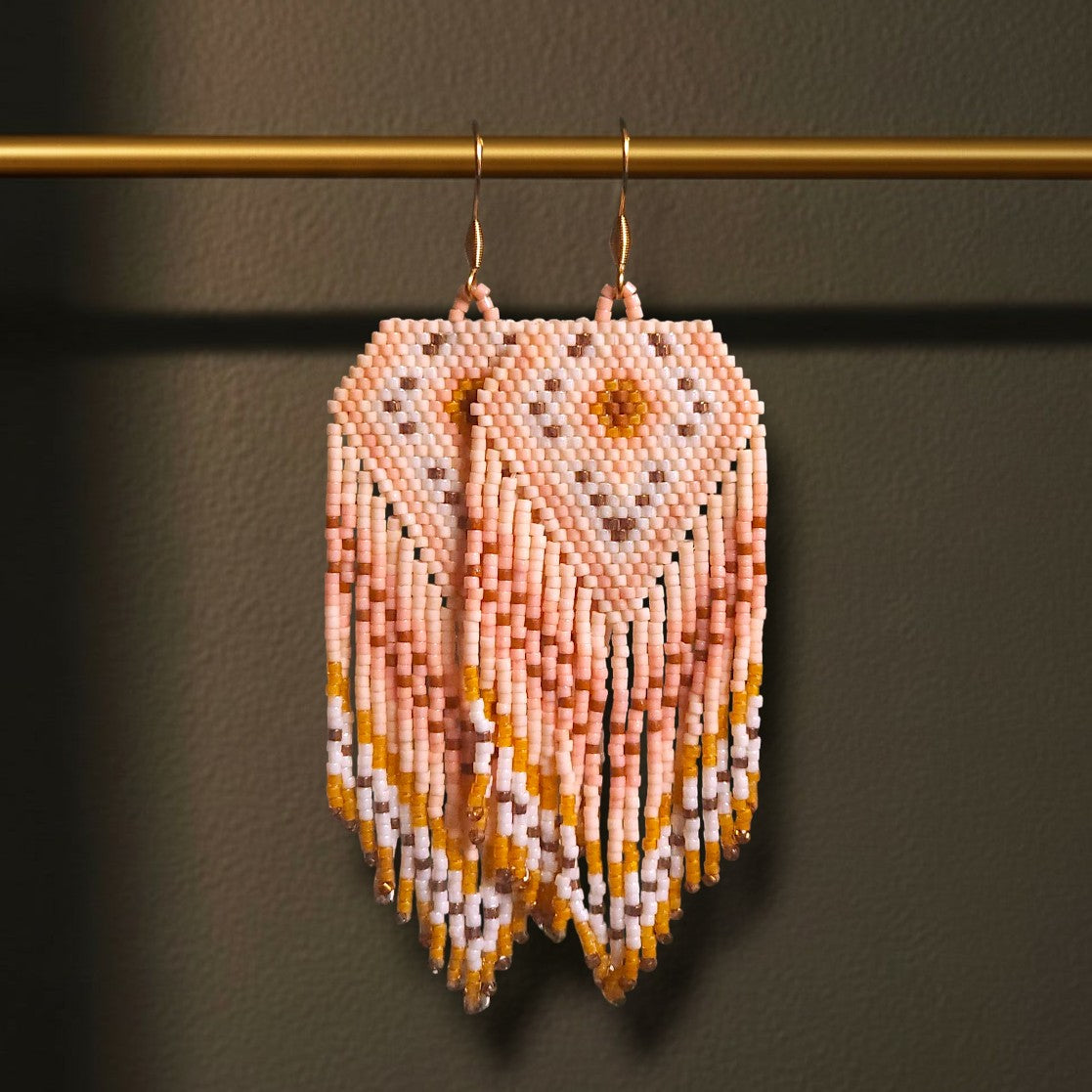 Handgewebte Perlenohrringe Bianco Pesca, aufgehängt an einer goldenen Präsentationsstange vor einem dunklen Hintergrund. Das Design zeigt ein helles Rautenmuster in Weiß, Apricot und Pfirsichtönen mit langen, fließenden Perlenfransen.