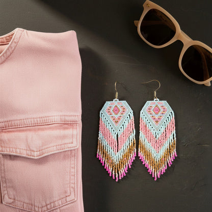 Auf einer dunklen, strukturierten Oberfläche liegen zwei lange Perlenohrringe mit Fransen im Zentrum. Das Design kombiniert hellblaue und rosa Pastelltöne mit erdigen Goldbraun-Nuancen und leuchtend pinken Spitzen. Links im Bild ist eine rosa Jeansjacke angeschnitten, rechts oben vervollständigt eine beige Sonnenbrille das modische Stillleben. Sanftes Licht setzt die feinen Details der handgewebten Schmuckstücke von EDELATELIER. in Szene.