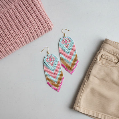 Ein hellgrauer Hintergrund zeigt ein flach liegendes Arrangement mit handgefertigten Fransenohrringen im Fokus. Die Ohrringe bestehen aus filigranen Glasperlen in den Farben Hellblau, Rosa, Goldbraun und kräftigem Pink, die ein geometrisches Rautenmuster und einen V-förmigen Farbverlauf bilden. Links oben liegt ein Teil eines altrosa Strickpullovers, rechts unten ist der Bund eines beigen Rocks zu sehen.