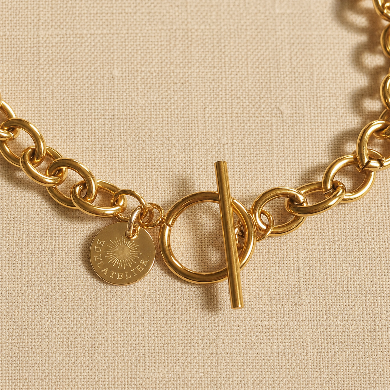 Detailaufnahme der Catena Luce Halskette aus 18 Karat PVD-vergoldetem Edelstahl mit T-Verschluss und EDELATELIER-Logoplättchen auf warmem hellbeigem Leinen.