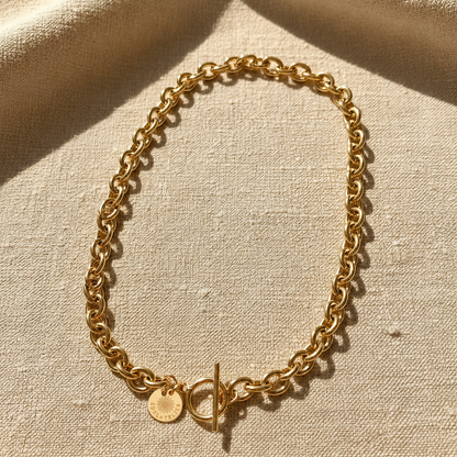Goldfarbene Catena Luce Halskette aus 18 Karat PVD-vergoldetem Edelstahl mit markanter Forçat-Gliederung und T-Verschluss auf warmem hellbeigem Leinen.