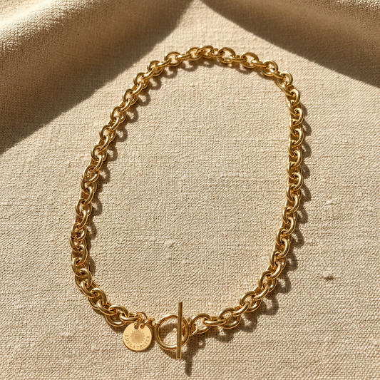 Goldfarbene Catena Luce Halskette aus 18 Karat PVD-vergoldetem Edelstahl mit markanter Forçat-Gliederung und T-Verschluss auf warmem hellbeigem Leinen.