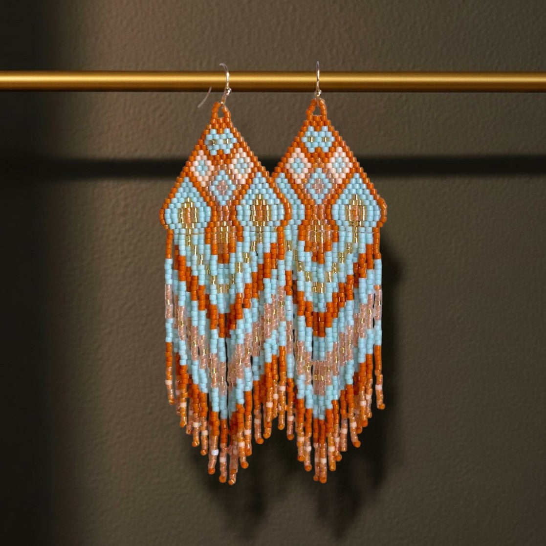 Handgewebte Miyuki Perlenohrringe Modell Celeste Mattone von EDELATELIER. dekorativ hängend an einer goldenen Metallstange. Das geometrische Muster in Hellblau, Terrakotta und Creme wird durch den dunklen Hintergrund bei einer Statement-Länge von 11 cm besonders betont.