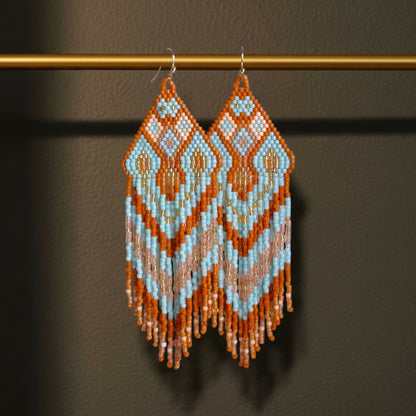 Handgewebte Miyuki Perlenohrringe Modell Celeste Mattone von EDELATELIER. dekorativ hängend an einer goldenen Metallstange. Das geometrische Muster in Hellblau, Terrakotta und Creme wird durch den dunklen Hintergrund bei einer Statement-Länge von 11 cm besonders betont.