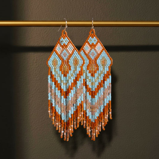 Handgewebte Miyuki Perlenohrringe Modell Celeste Mattone von EDELATELIER. dekorativ hängend an einer goldenen Metallstange. Das geometrische Muster in Hellblau, Terrakotta und Creme wird durch den dunklen Hintergrund bei einer Statement-Länge von 11 cm besonders betont.