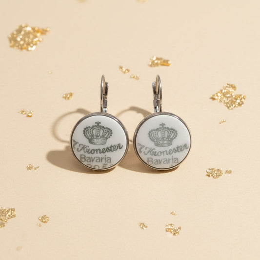 Handgemachte Porzellanohrringe Coronula aus weißem Upcycling-Porzellan mit grün-grauem Vintage-Stempel einer Krone und Kronester Bavaria Schriftzug, liegend auf beigem Hintergrund mit Goldflocken von EDELATELIER.