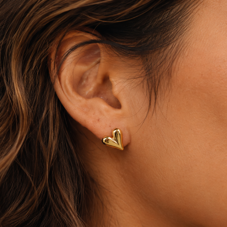 Goldene Herz-Ohrstecker Cuore Piccolo am Ohr eines Models mit braunen Wellen und warmer Haut, Nahaufnahme.