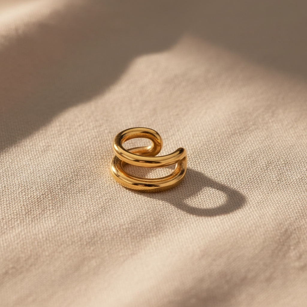 Goldener Doppio Ear Cuff aus Edelstahl auf warmen Leinenstoff.