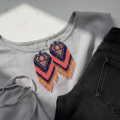 Handgemachte Perlenohrringe Eclissi Dorata von EDELATELIER. Lange Franseneohrringe mit geometrischem Rautenmuster in Dunkelblau, Koralle, Grau und Gold, stilvoll arrangiert auf einer hellgrauen Bluse und einer schwarzen Jeans.