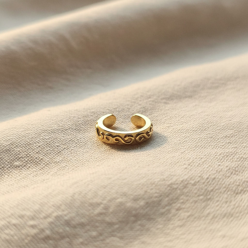 Zierlicher goldfarbener Arabesco Ear Cuff aus Edelstahl mit geschwungenem Reliefmuster auf hellem Leinen.