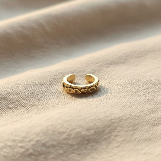 Zierlicher goldfarbener Arabesco Ear Cuff aus Edelstahl mit geschwungenem Reliefmuster auf hellem Leinen.