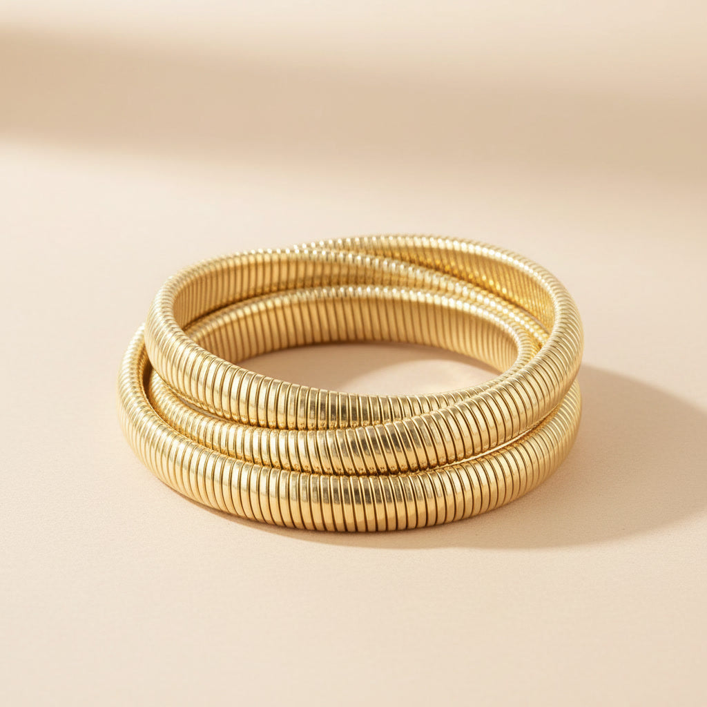 Goldfarbenes Spirale d’Oro Armband mit dreifacher Wickel-Optik und gerippter Struktur auf warmem beigem Hintergrund.