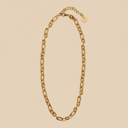 Goldfarbene Catena Halskette mit länglichen Gliedern, Karabiner und Verlängerungskette auf warmem cremefarbenem Hintergrund.