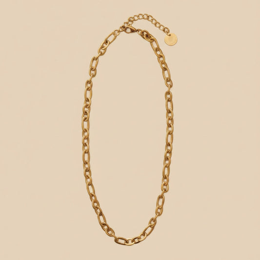Goldfarbene Catena Halskette mit länglichen Gliedern, Karabiner und Verlängerungskette auf warmem cremefarbenem Hintergrund.