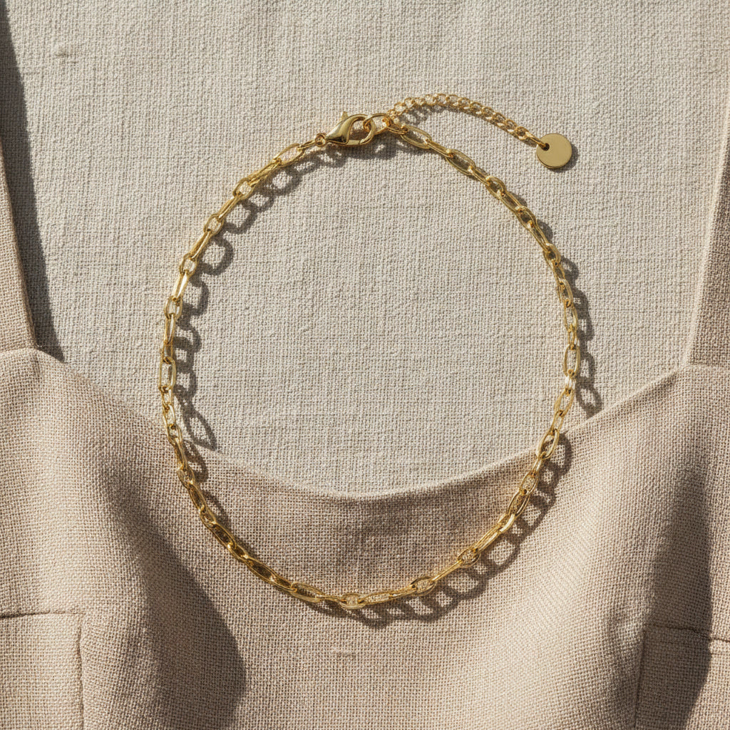 Goldfarbene Catena Halskette mit länglichen Gliedern auf hellem Leinenkleid im Sonnenlicht.