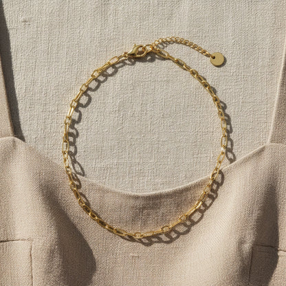 Goldfarbene Catena Halskette mit länglichen Gliedern auf hellem Leinenkleid im Sonnenlicht.