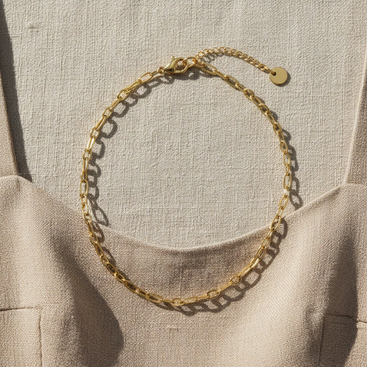 Goldfarbene Catena Halskette mit länglichen Gliedern auf hellem Leinenkleid im Sonnenlicht.
