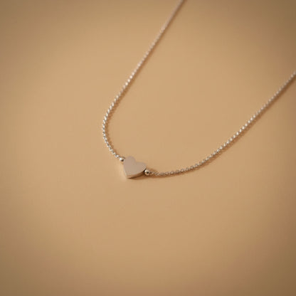 Detailaufnahme der silberfarbenen Cuore Halskette mit kleinem Herzanhänger auf warmem beigem Hintergrund.