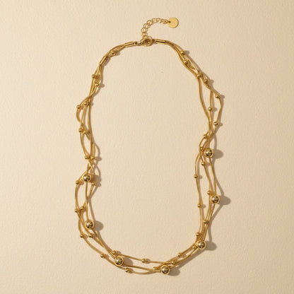 Mehrreihige goldfarbene Edelstahl-Halskette mit feinen Kugeldetails auf warmem cremefarbenem Untergrund.