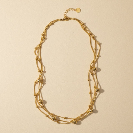 Mehrreihige goldfarbene Edelstahl-Halskette mit feinen Kugeldetails auf warmem cremefarbenem Untergrund.